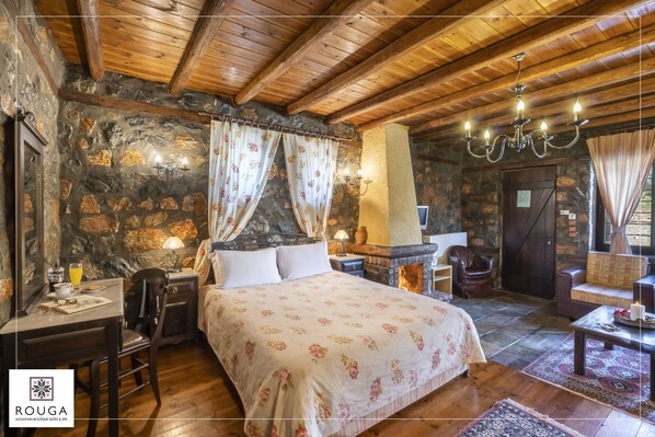 A cozy room with a stone wall, wooden ceiling, a bed with floral bedding, a desk with a lamp, and a chandelier.