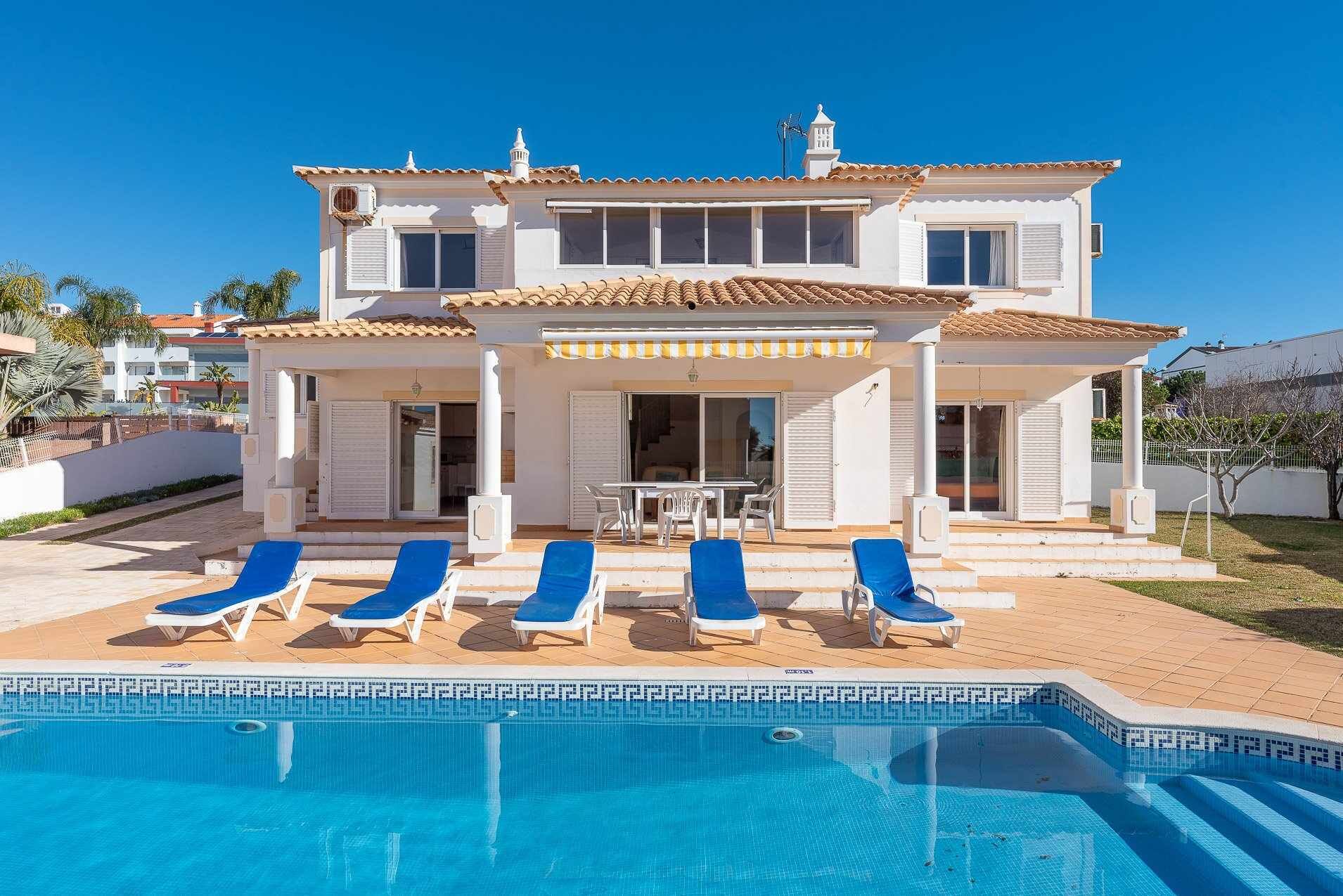 Een tweelaags huis met een zwembad, een witte gevel en een overdekt terras met blauwe lounge fauteuils.