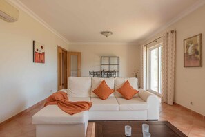 A living room with a white sofa, orange cushions, a coffee table with glasses, a dining area with a table and chairs, and a window with curtains. - Villa Costa by Gabi Miguel (Sesmarias)