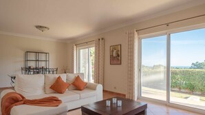 A spacious living room with a white sofa, orange cushions, a coffee table, and large windows offering a view of greenery and a distant landscape. - Villa Costa by Gabi Miguel (Sesmarias)