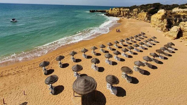 A beach with rows of sun umbrellas and lounge chairs, a clear blue sky, and a rocky coastline. - Villa Costa by Gabi Miguel (Sesmarias)