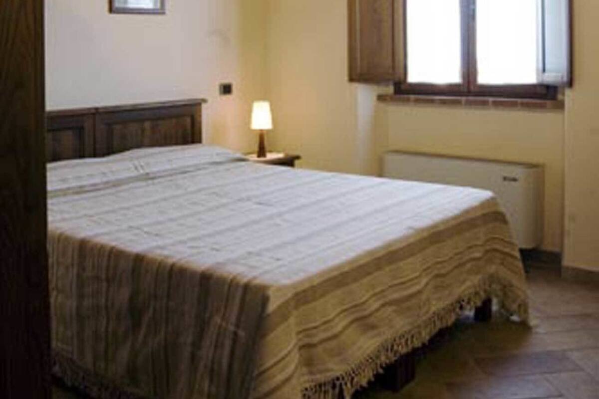 Quarto com teto de madeira, cama com colcha branca, mesa de cabeceira com abajur, janela com venezianas de madeira e radiador abaixo da janela.