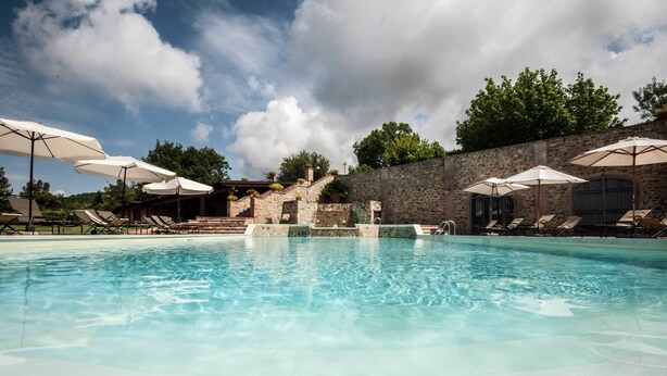 Piscina com espreguiçadeiras e guarda-sóis, cercada por um muro de pedra e árvores.