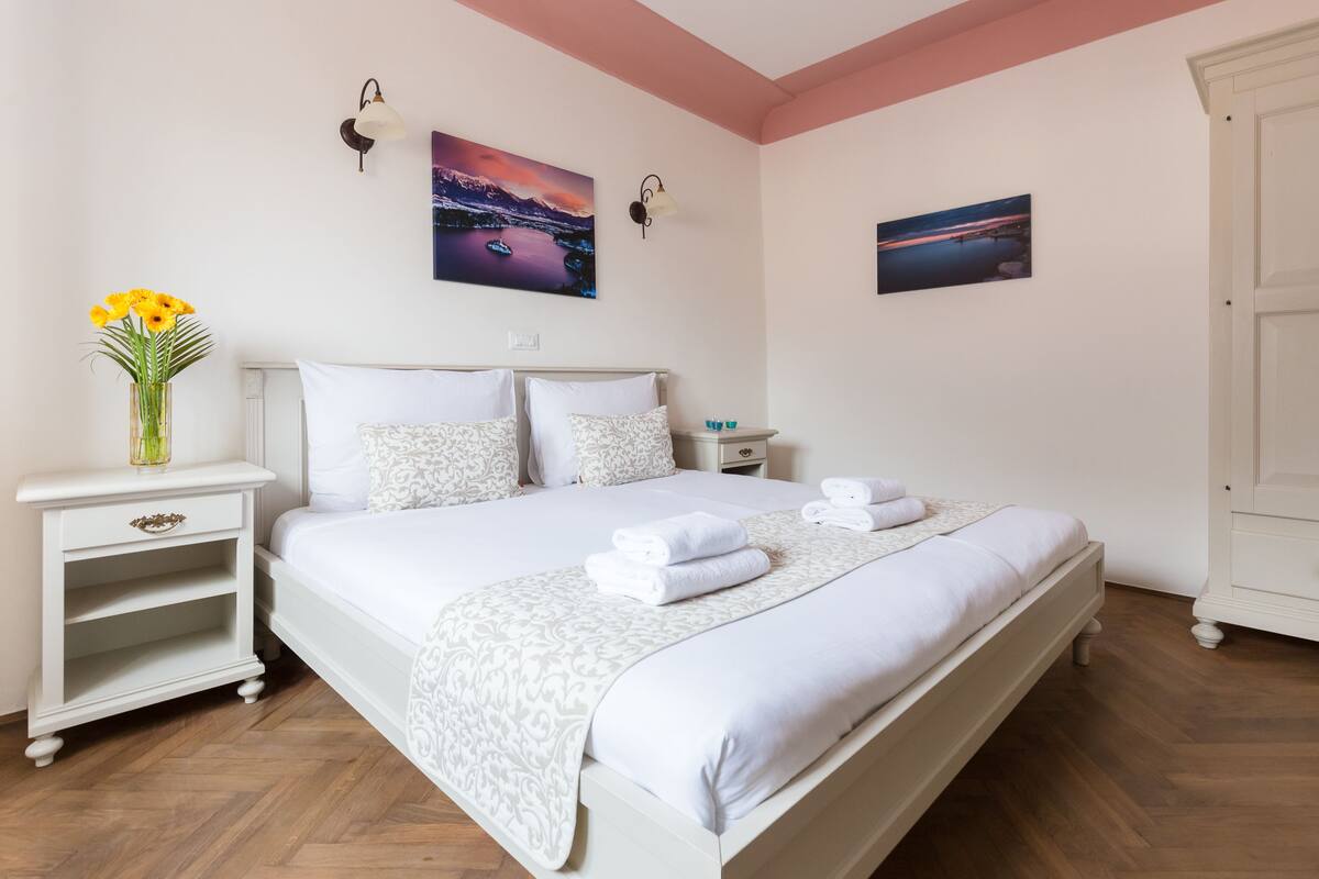 A neatly made bed with white linens and a decorative throw, a bedside table with a vase of flowers, and two wall-mounted lamps.