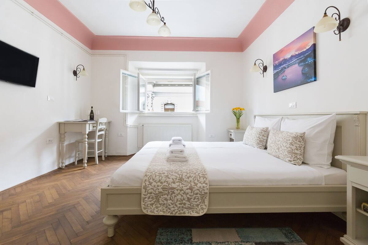 A bedroom with a bed, a desk, a chair, a window, and a painting on the wall.
