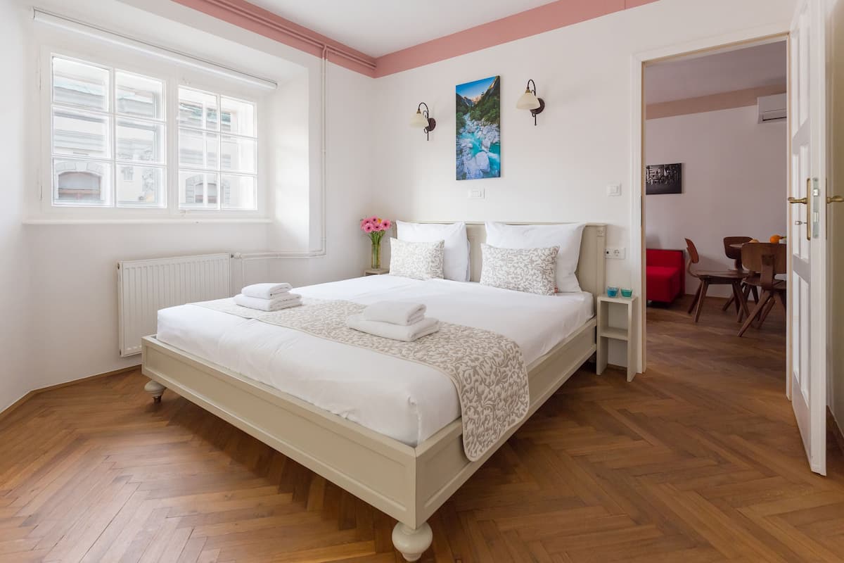 A bedroom with a large bed, a window, a painting, and a view into another room.