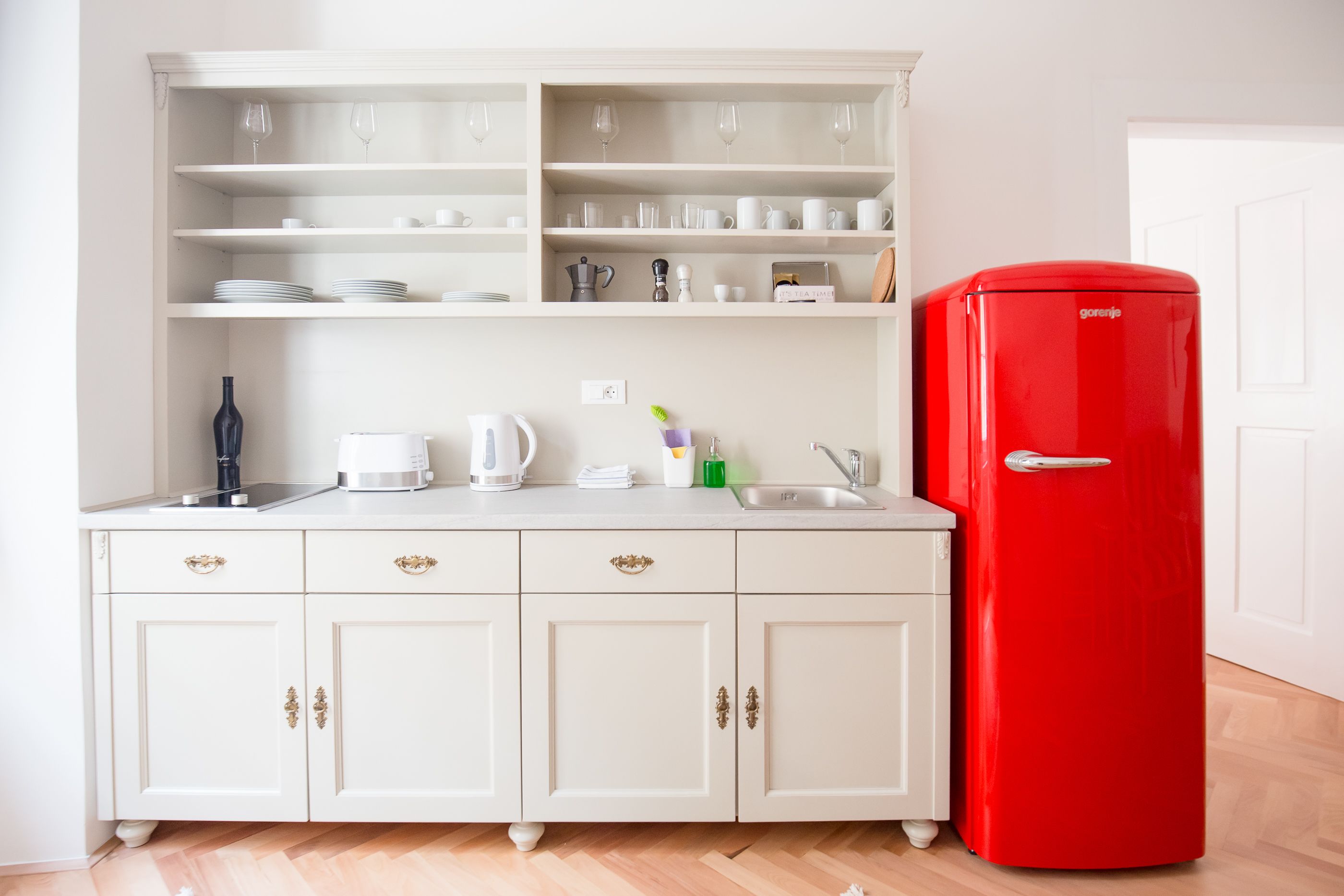 Una cocina con armarios blancos, un refrigerador rojo y estantes con vasos y vajilla.