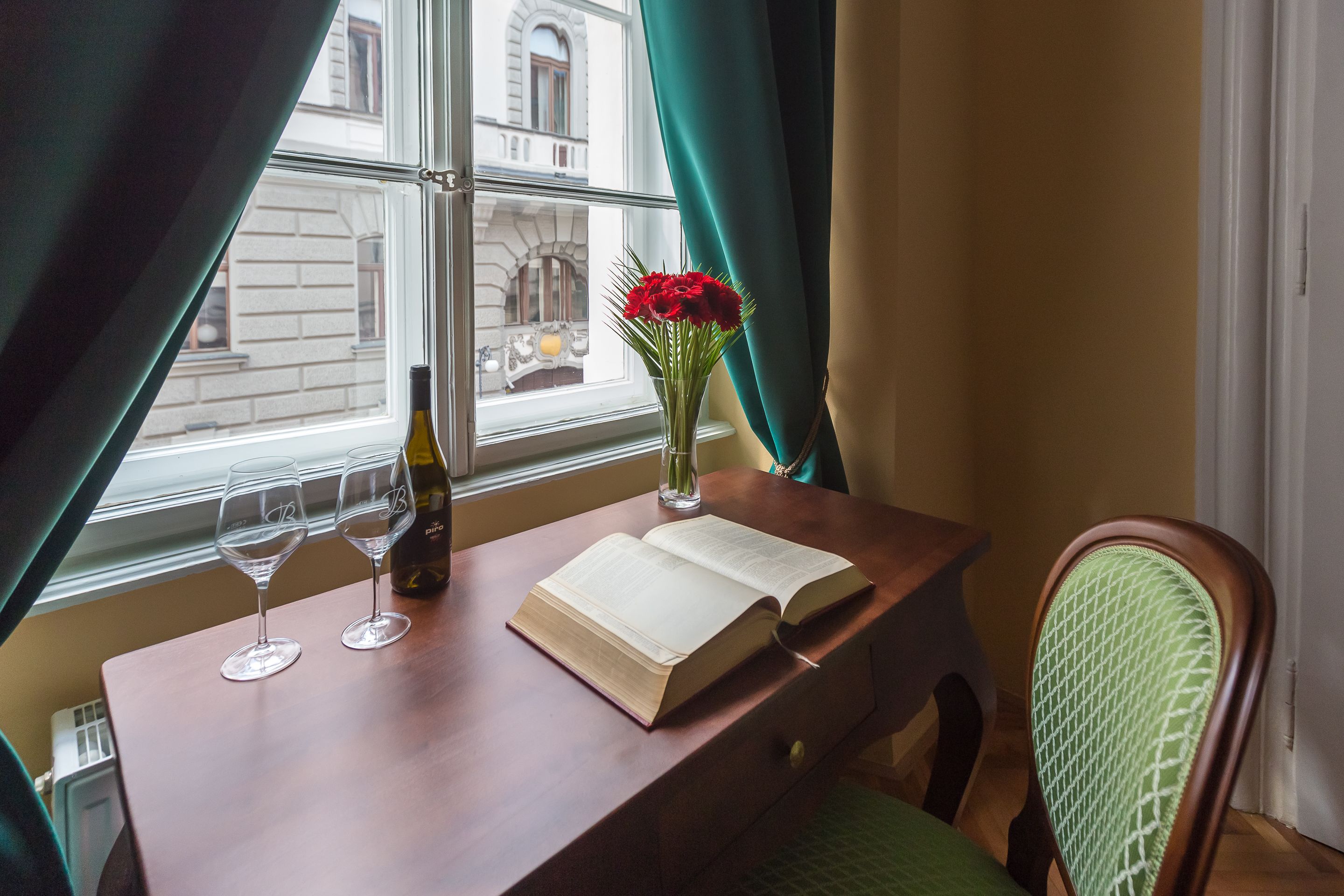 Un bureau en bois sur lequel se trouve un livre ouvert, deux verres à vin et une bouteille de vin. Un vase rempli de fleurs rouges est posé sur le bureau.