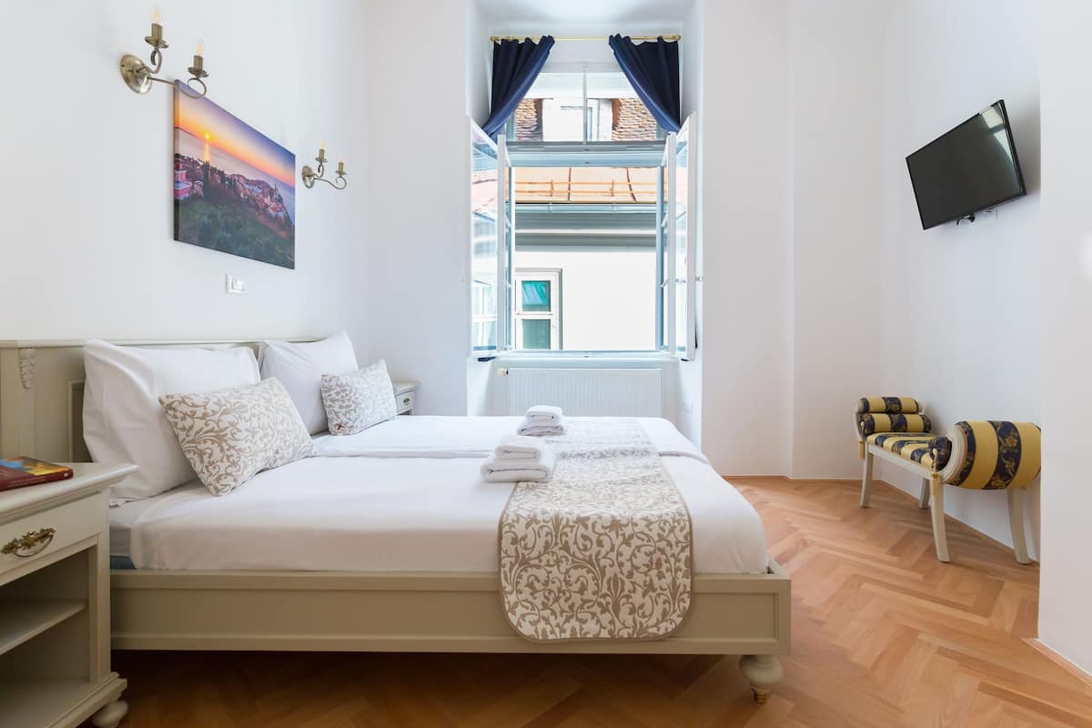 A bedroom with a bed, a nightstand, a TV, a window with curtains, and a view of the outside.