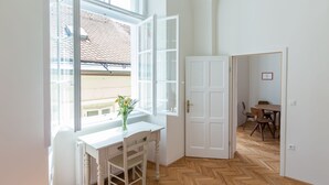A room with a white desk, a chair, a window with a view of a building, and a wooden herringbone floor. - Barbo Palace (Ljubljana)