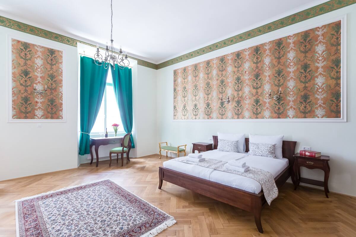 A bedroom with a wooden bed, a desk with a chair, a small table with a chair, and a large window with curtains.