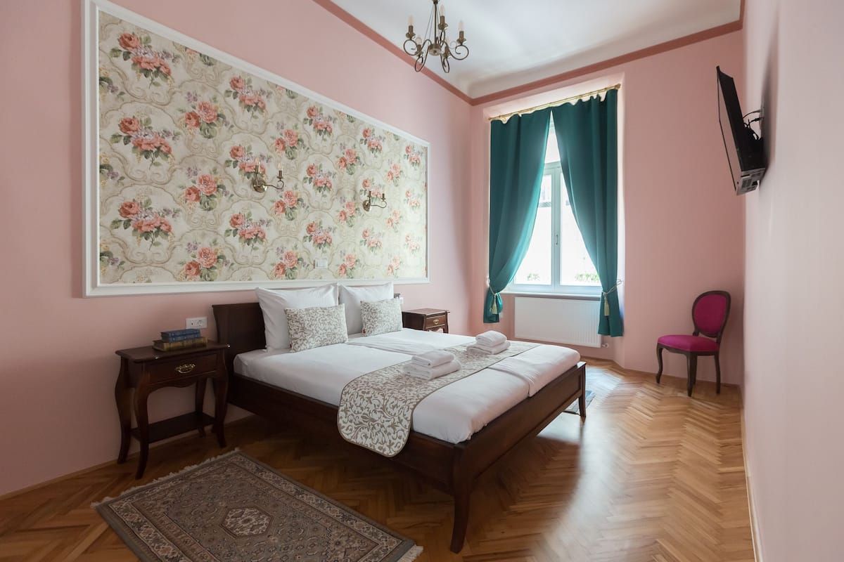 A bedroom with a large floral wallpaper, a wooden bed, a nightstand, a chair, and a TV mounted on the wall.