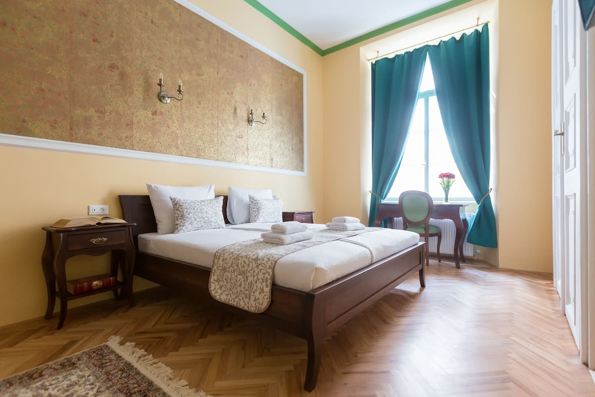 A neatly arranged bedroom with a large bed, a bedside table with a book, a chair, and a window with curtains.