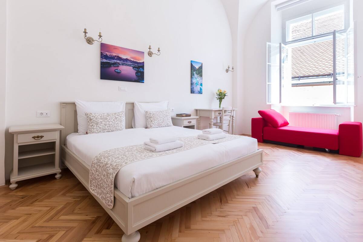 A spacious bedroom with a large bed, a red sofa, and a view of the city through a large window.