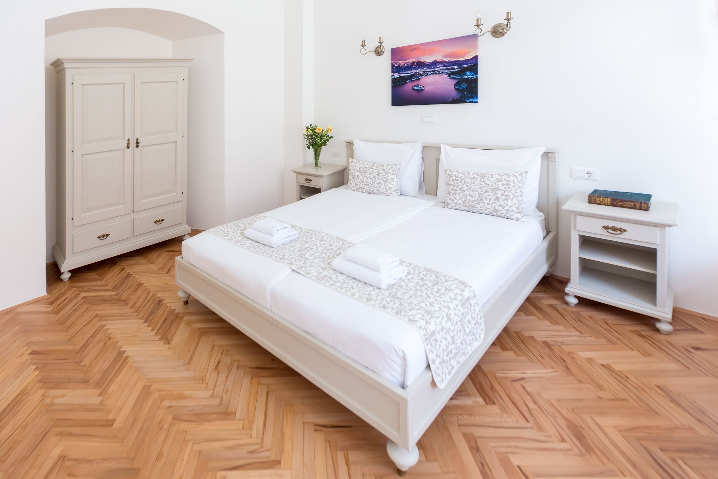 Una camera da letto ordinata con un letto grande, un armadio, un comodino con un vaso di fiori e un quadro appeso al muro.