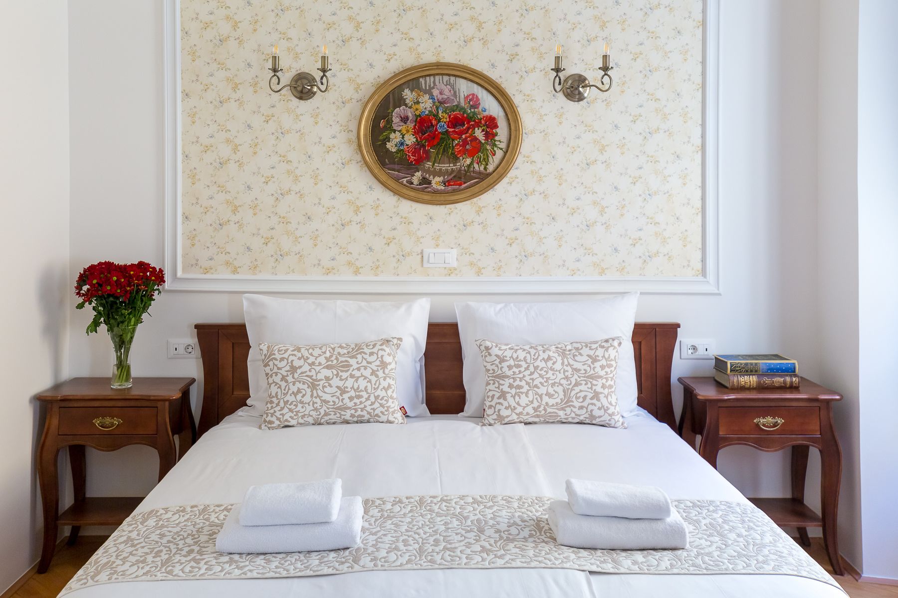 Un lit bien fait, avec du linge de lit blanc et des coussins décoratifs, un vase de fleurs rouges sur une table de chevet, et une œuvre d’art florale encadrée au-dessus du lit.