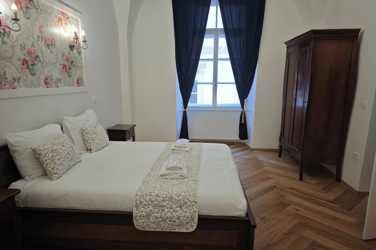 A neatly made bed with a patterned bedspread and pillows, a wooden floor, a dark wooden wardrobe, and a window with blue curtains.