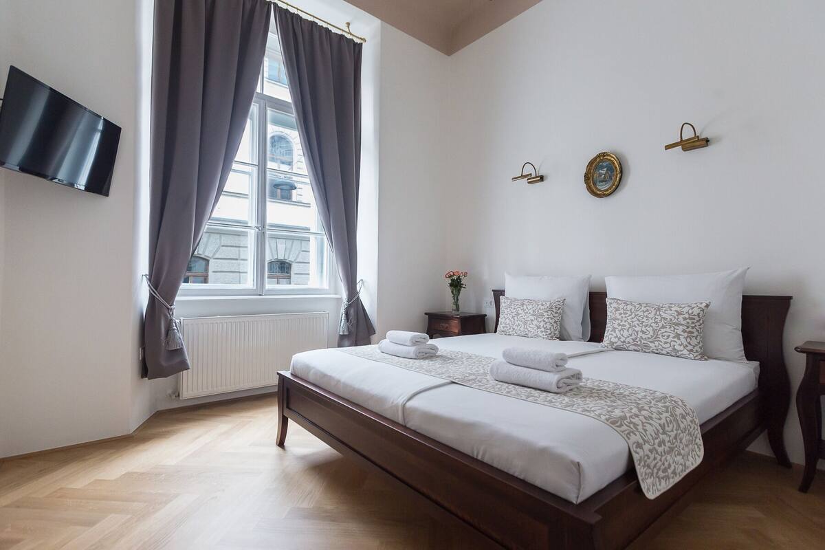 A bedroom with a large bed, a TV mounted on the wall, and a window with curtains.
