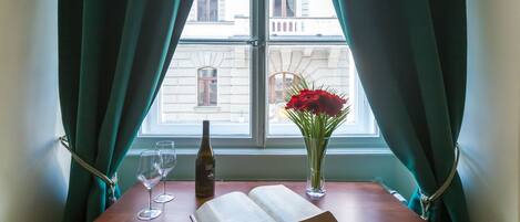 Una mesa de madera con un libro abierto, dos copas de vino, una botella y un florero con flores junto a una ventana con cortinas.