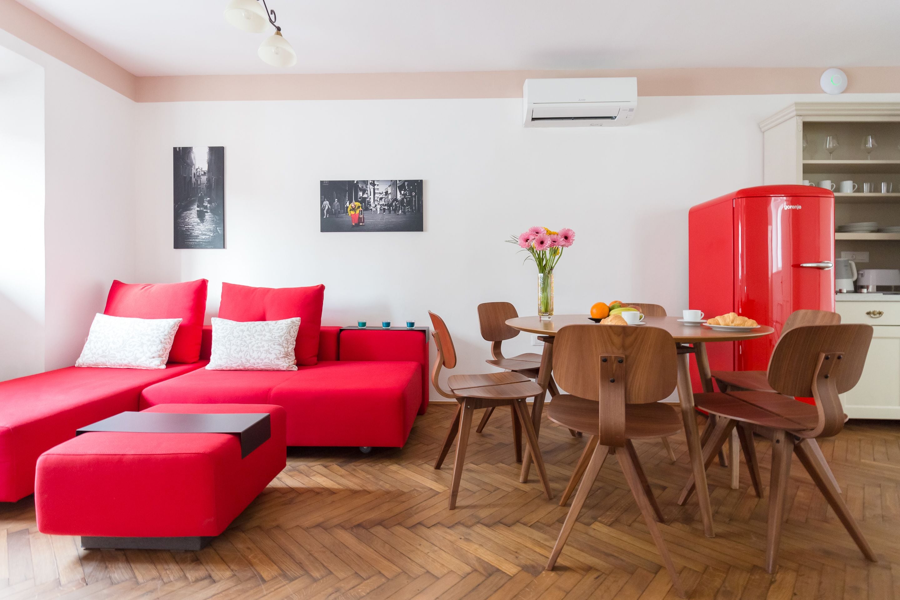 A modern living room with a red sofa, wooden dining table, and chairs. There is a kitchen area with a red refrigerator and a white cabinet.