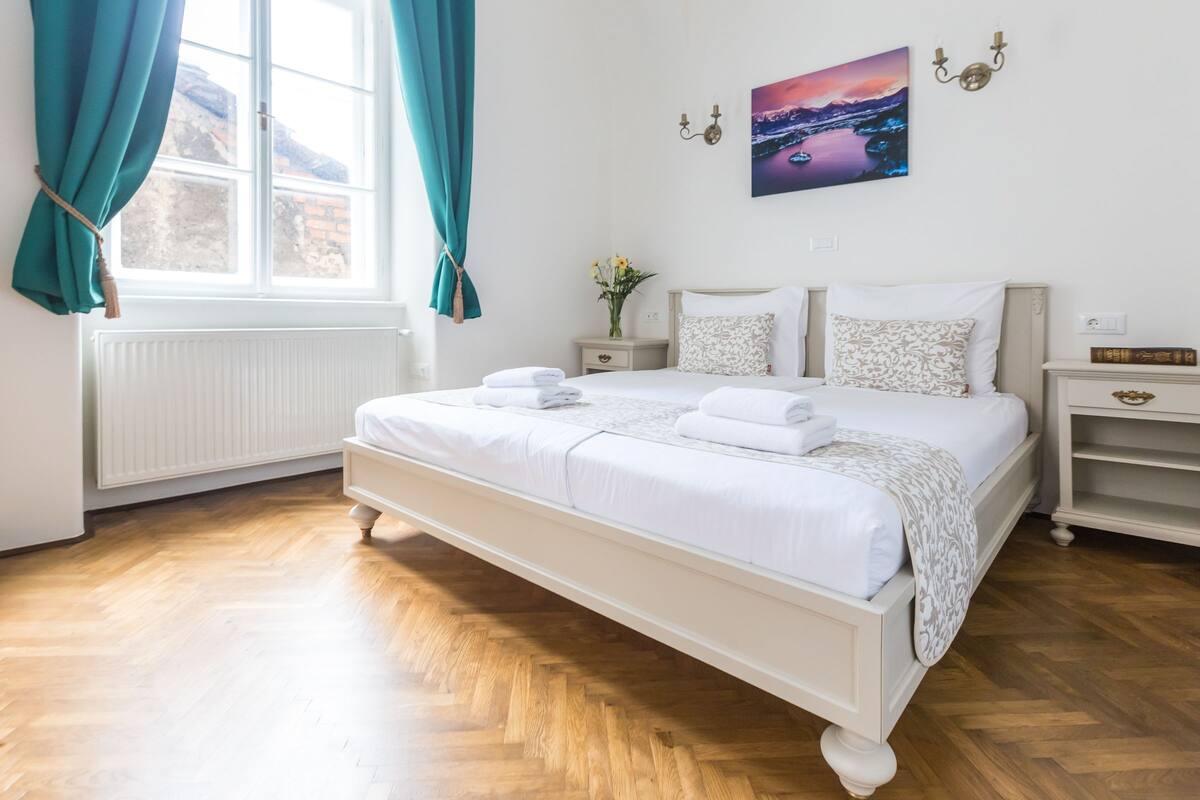 A neatly arranged bedroom with a large bed, a bedside table, a nightstand, and a wall-mounted lamp.