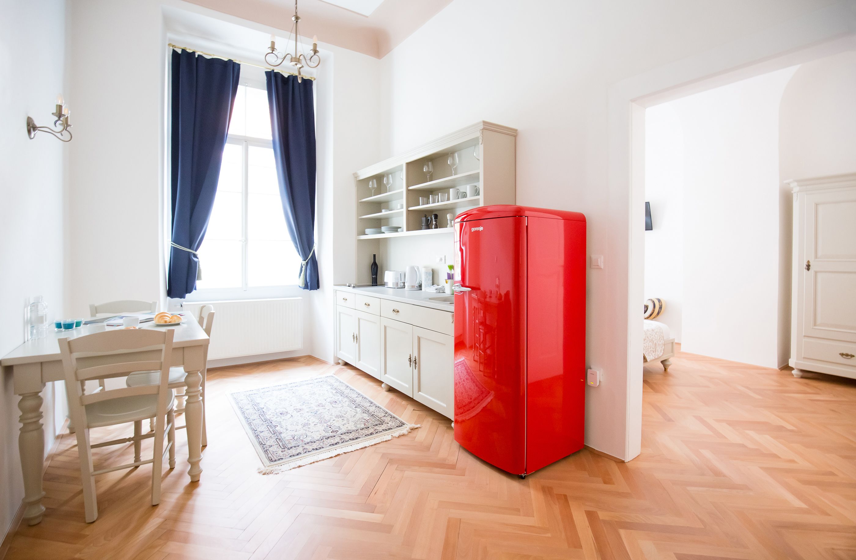 Une petite cuisine avec un meuble blanc, un réfrigérateur rouge, une table à manger avec des chaises et une fenêtre ornée de rideaux bleus.