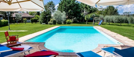 A rectangular swimming pool with a tiled edge, surrounded by a brick patio. There are blue lounge chairs with red towels, and a white umbrella structure overhead.