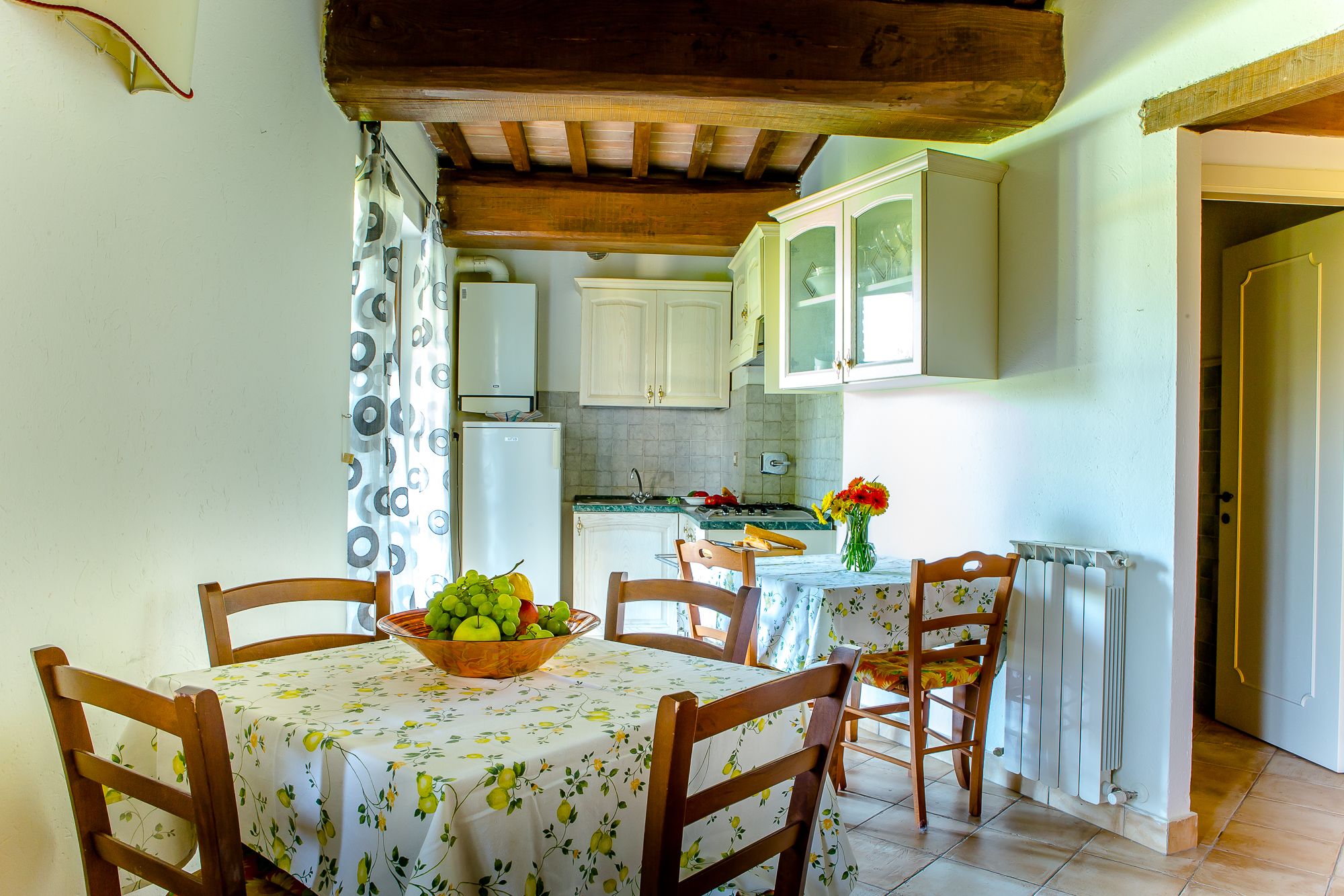 A dining area with a table set for two, a bowl of fruit, and a vase of flowers.