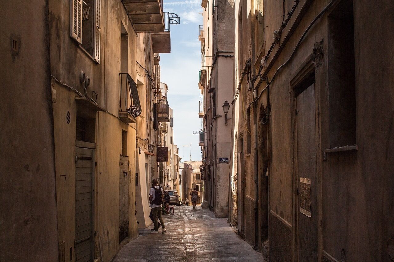 A narrow, sunlit street flanked by old buildings with balconies and shutters.