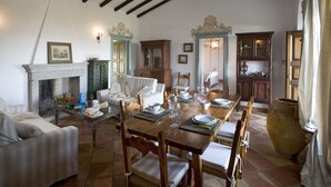 A dining room with a wooden table set for four, surrounded by wooden chairs. There is a fireplace with a mantel, a blue vase, and a framed picture above it. - Villa Cannigione (Palau)