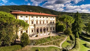 Un grand bâtiment à deux étages, surmonté d’un toit recouvert de tuiles rouges, entouré d’une végétation luxuriante et adossé à un paysage montagneux.
