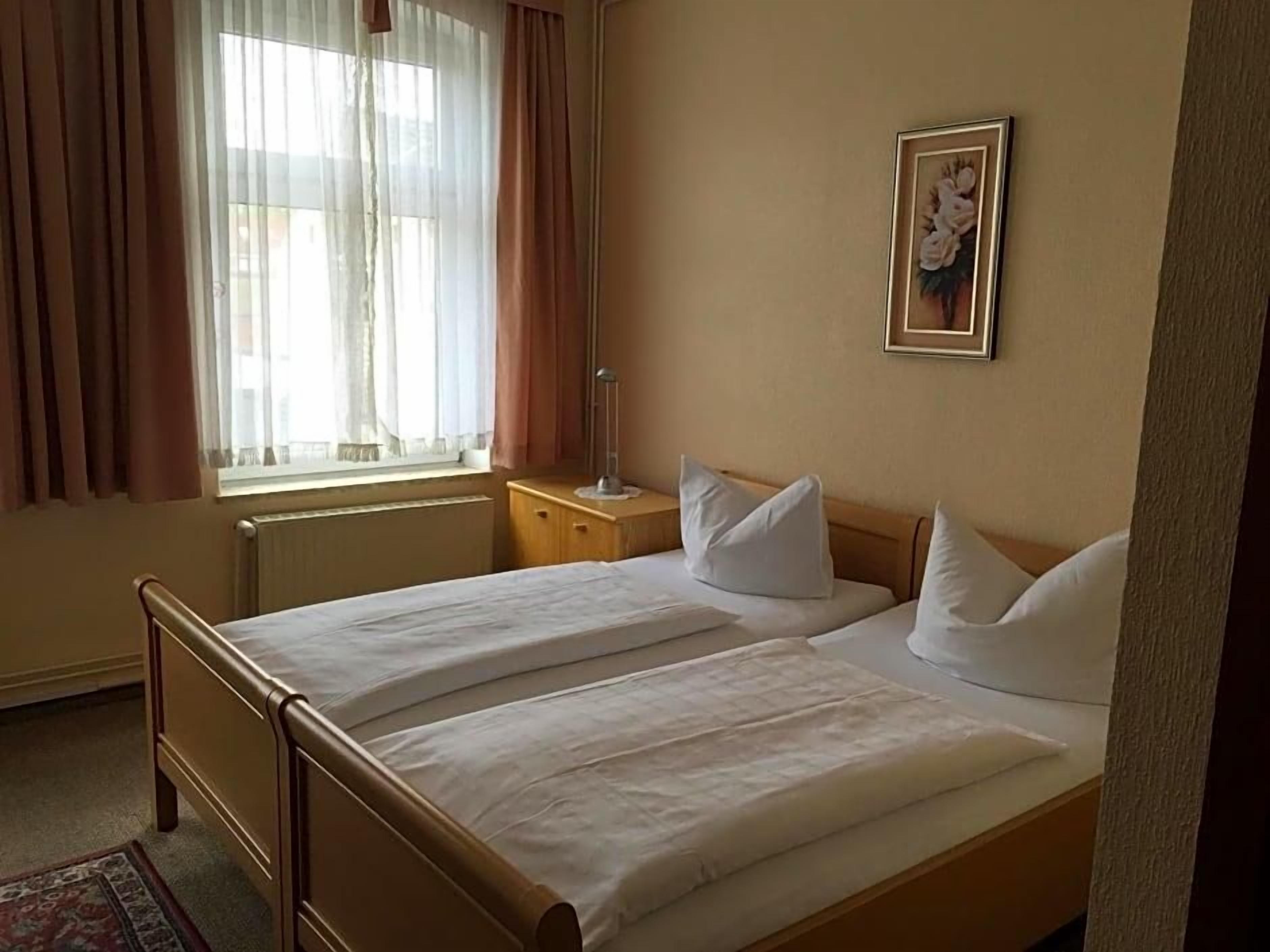 Ein Hotelzimmer mit zwei Betten, einem Nachttisch, einer Lampe, einem Fenster mit Vorhängen und einem gerahmten Bild an der Wand.