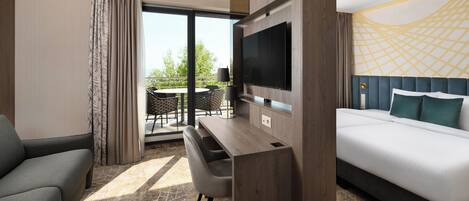 A hotel room with a bed, a TV mounted on the wall, a desk with a chair, and a view of the outdoors.