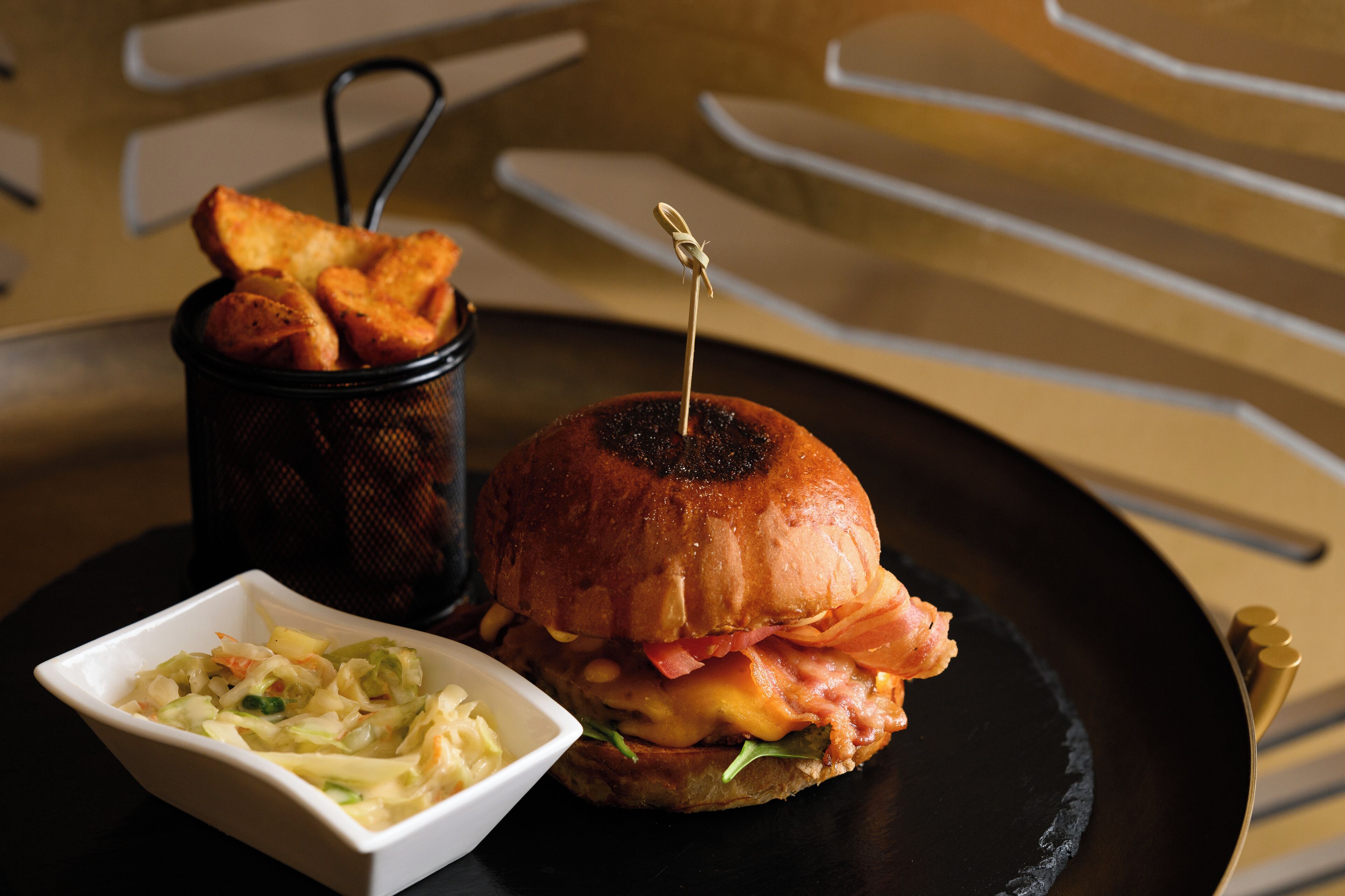 Hamburguesa con un palillo encima, servida en un plato negro acompañada de ensalada de col y patatas fritas.