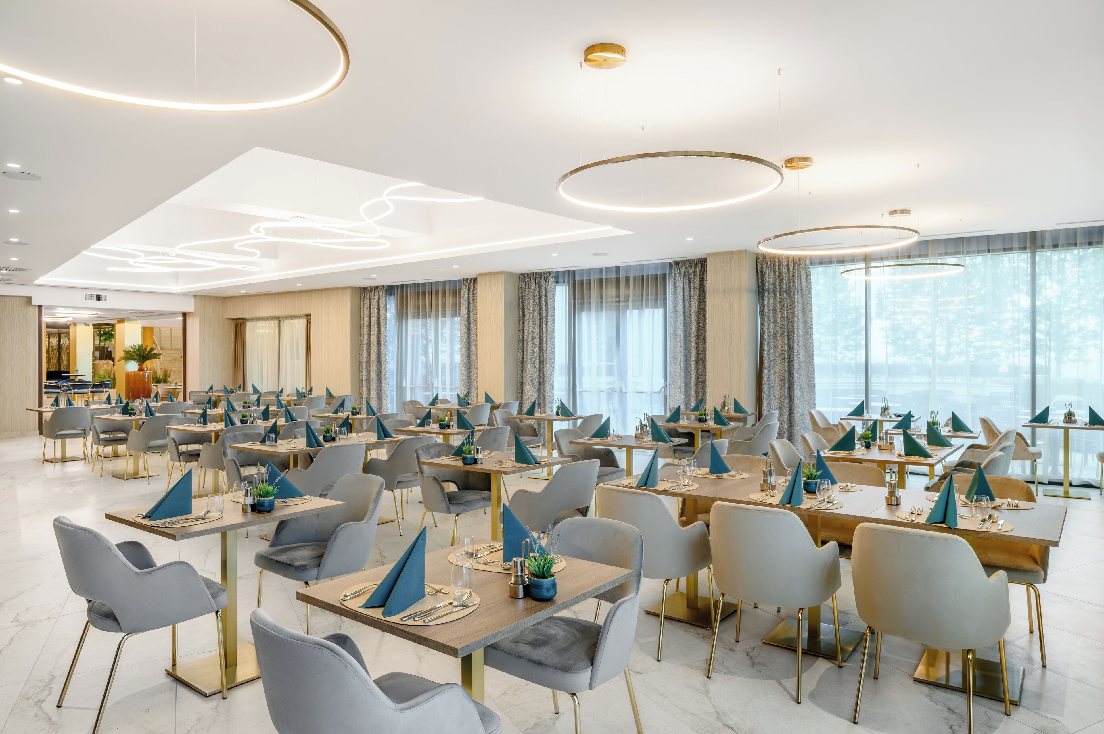A modern dining area with multiple tables and chairs, large windows, and contemporary lighting fixtures.
