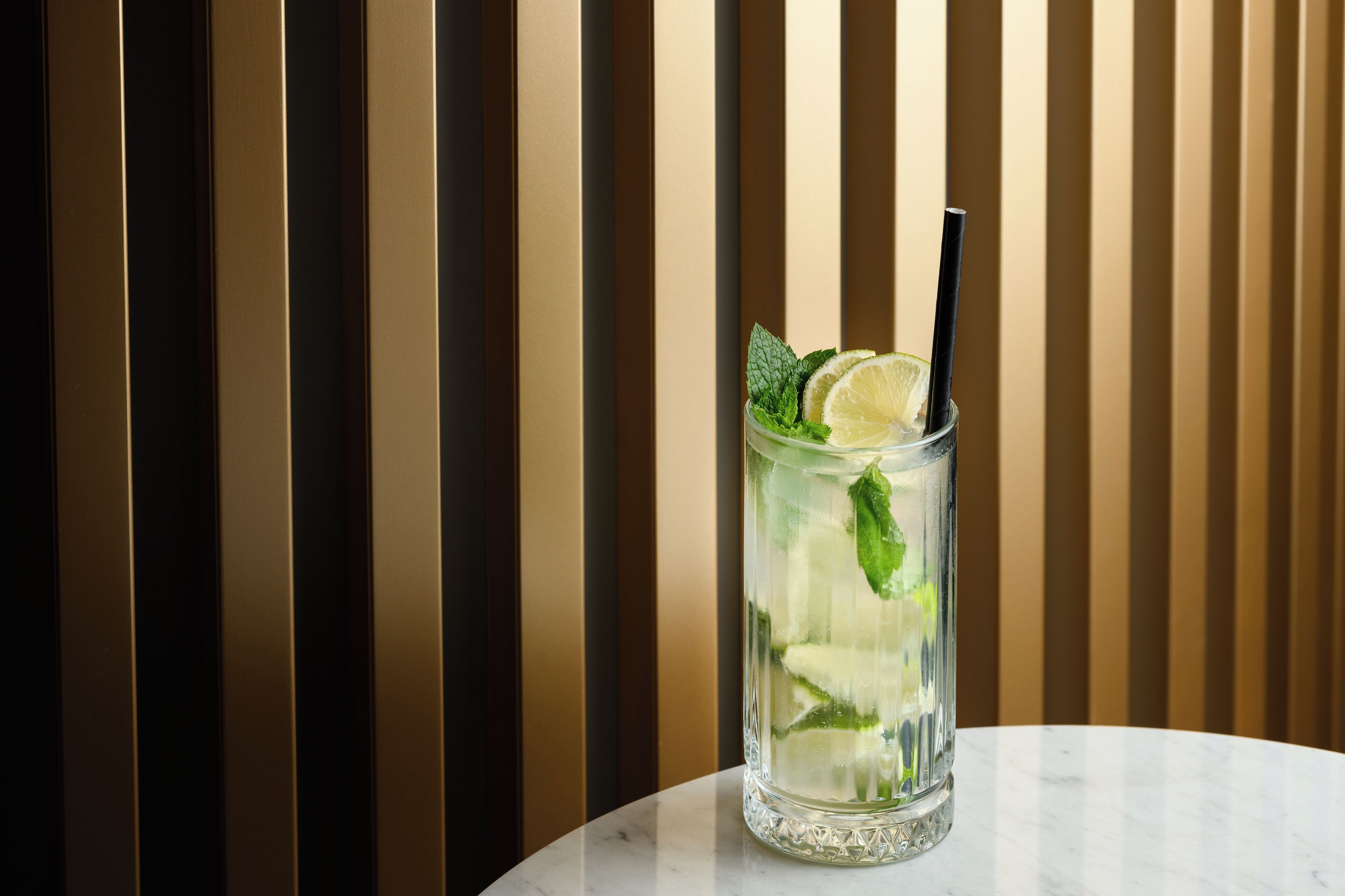 A clear glass with a straw, lime slice, and mint leaves on a marble surface against a striped background.