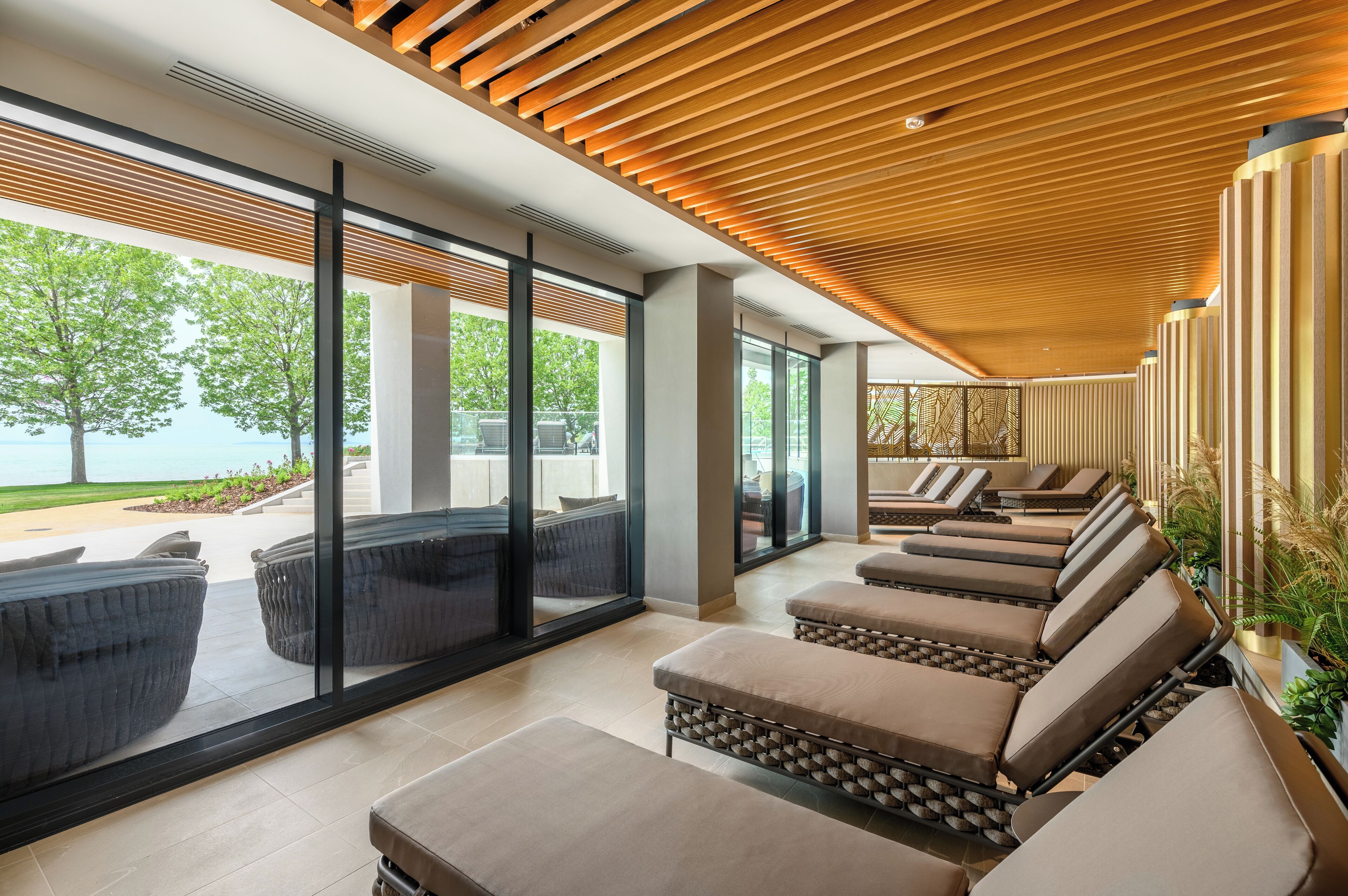 A modern lounge area with large windows, wooden ceiling slats, and outdoor seating by a pool.