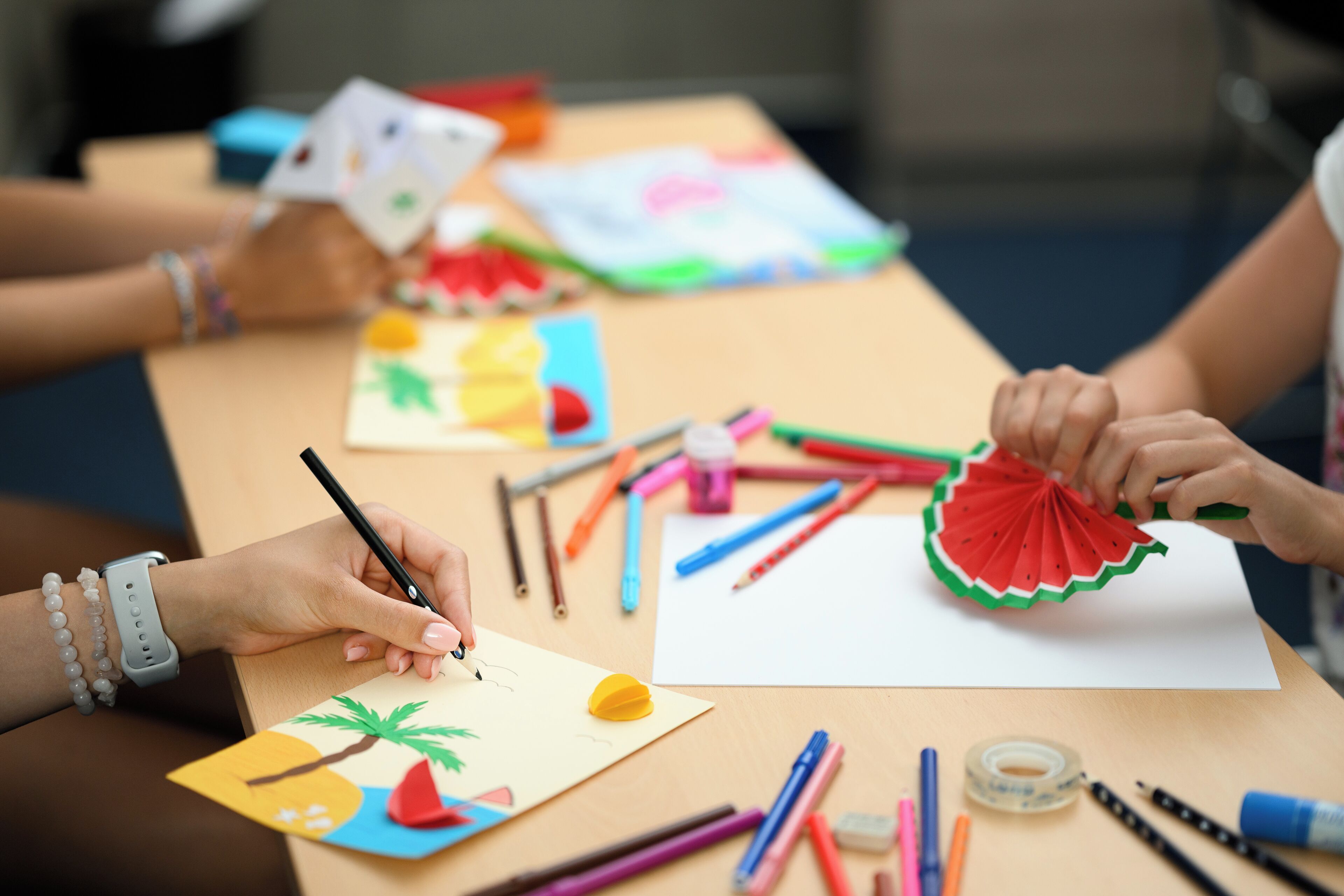 A table with various art supplies and a person drawing on a piece of paper.