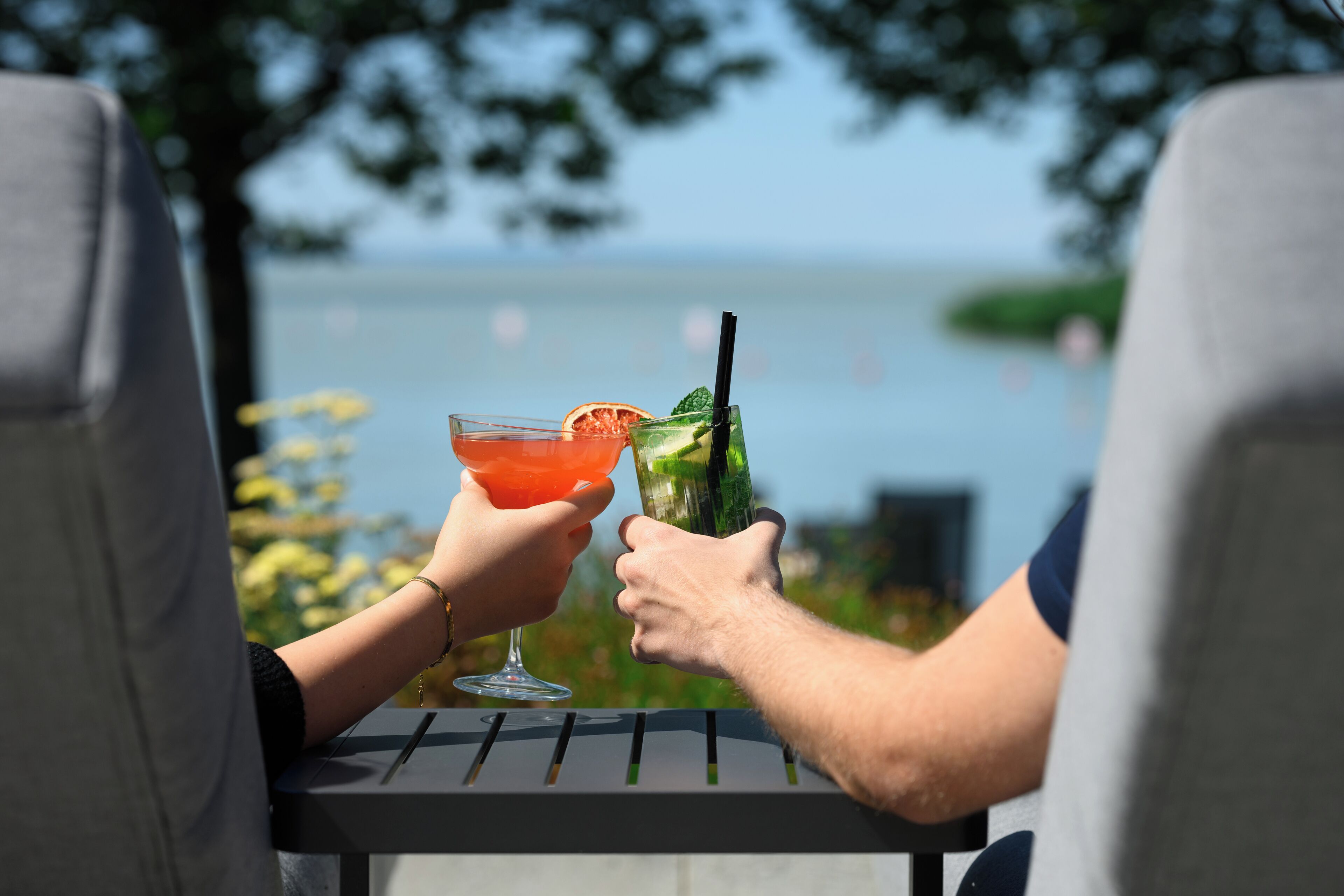 Two people sitting at a table with drinks, overlooking a body of water.