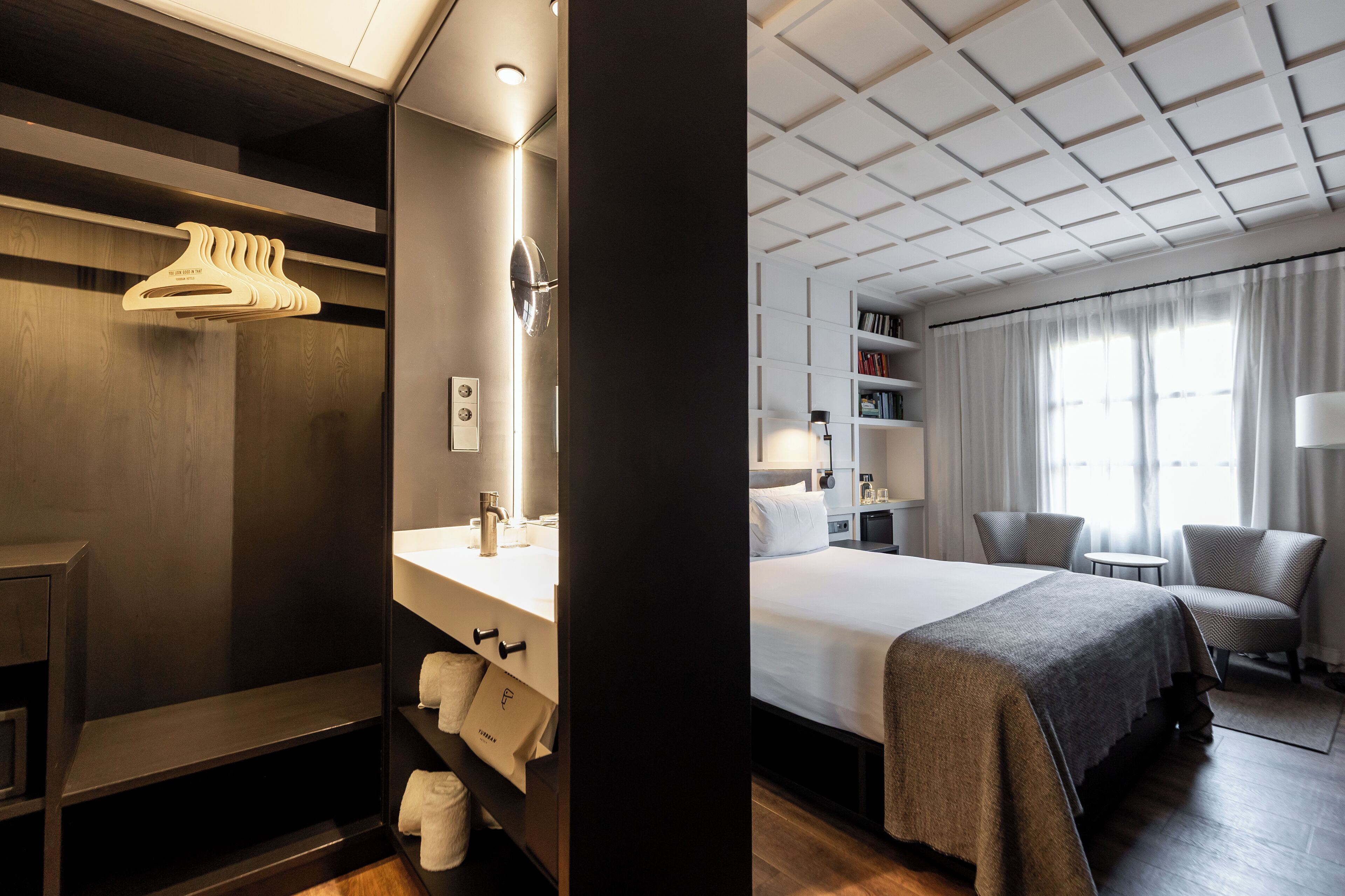 Habitación de hotel con cama, tocador con espejo, estante con toallas, perchero y ventana con cortinas.