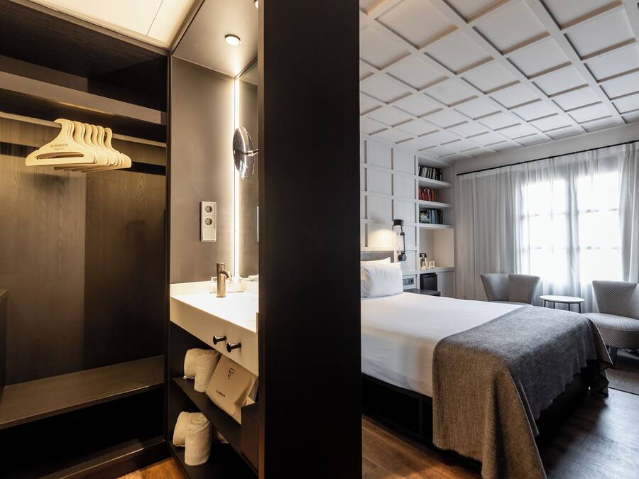 Habitación de hotel con cama, tocador con espejo, estante con toallas, perchero y ventana con cortinas.
