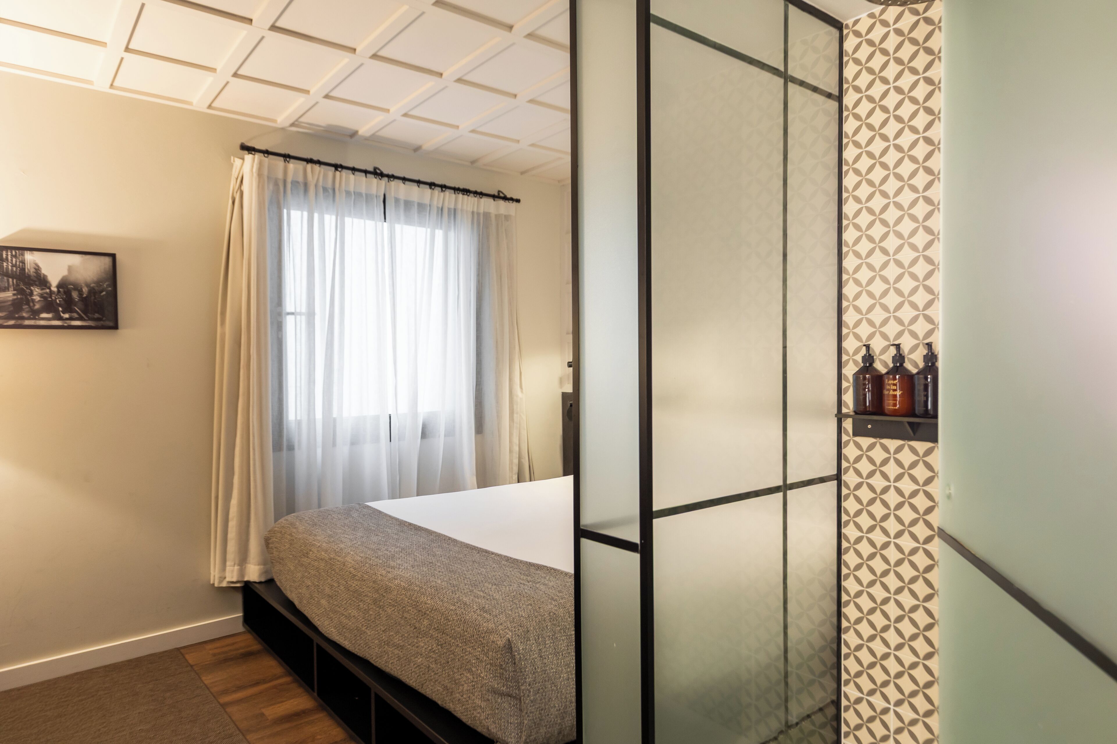 Ein Zimmer mit Bett, einem Fenster mit durchsichtigen Vorhängen, einem Bild an der Wand und einem gemusterten Wandbelag.