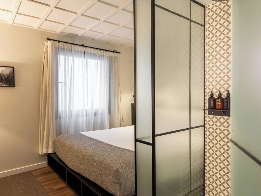 Ein Zimmer mit Bett, einem Fenster mit durchsichtigen Vorhängen, einem Bild an der Wand und einem gemusterten Wandbelag.
