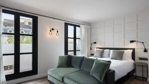 Une chambre d’hôtel moderne avec un grand lit, un canapé vert et une vue dégagée sur l’extérieur grâce à une grande fenêtre.