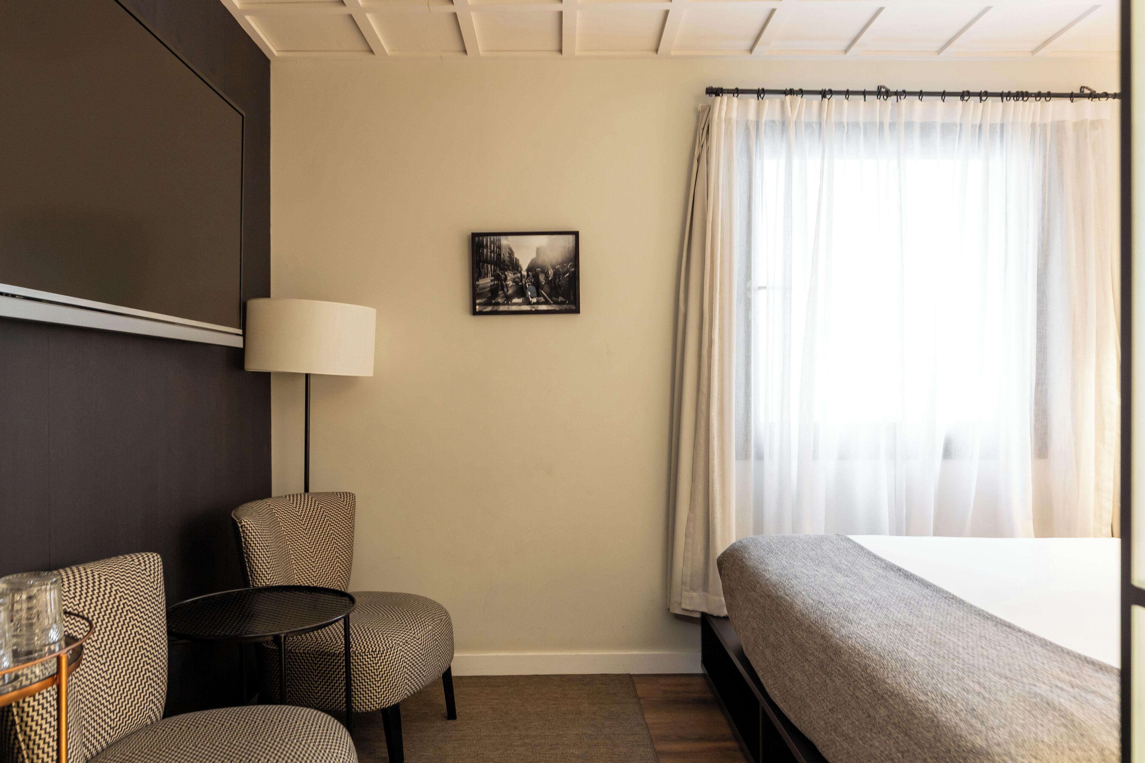 Quarto de hotel com cama, cadeira, mesinha, luminária e um quadro na parede.