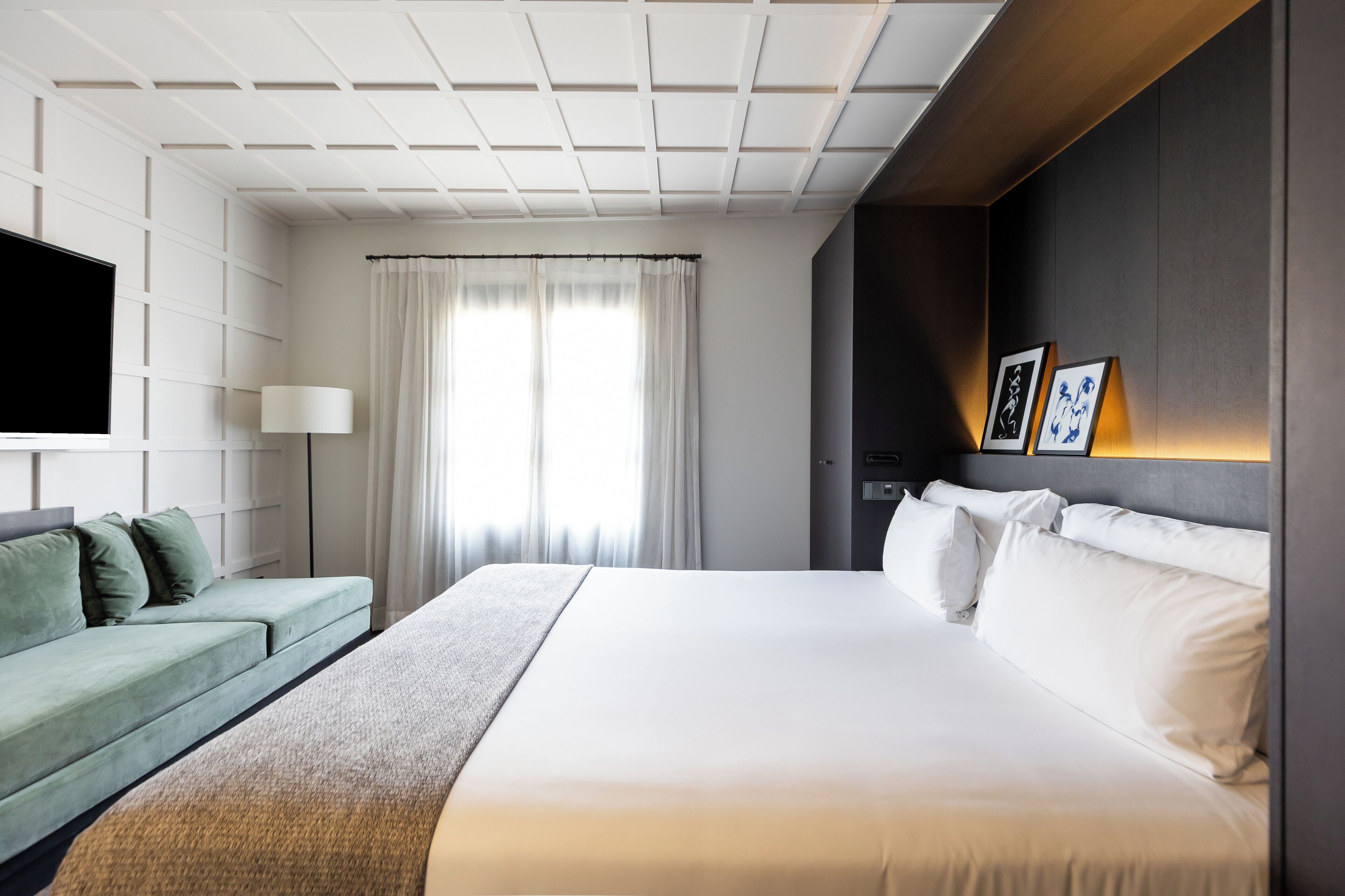 A modern hotel room with a large bed, a green sofa, a flat-screen TV, and a wall-mounted lamp.