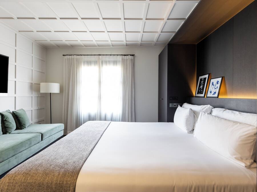 A modern hotel room with a large bed, a green sofa, a flat-screen TV, and a wall-mounted lamp.