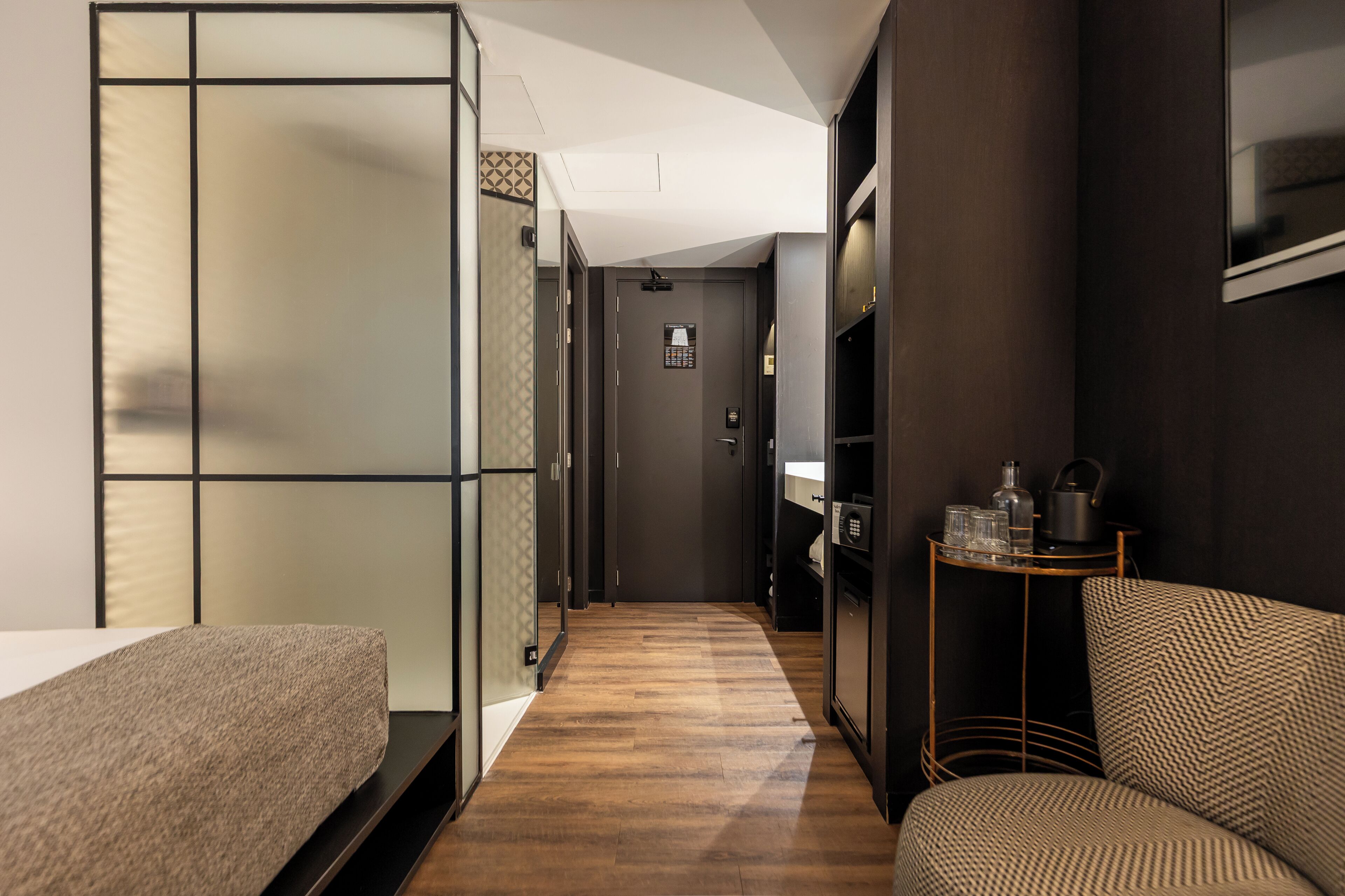 A narrow hallway with a bed on the left, a seating area with a chair on the right, and a small table with items on it.