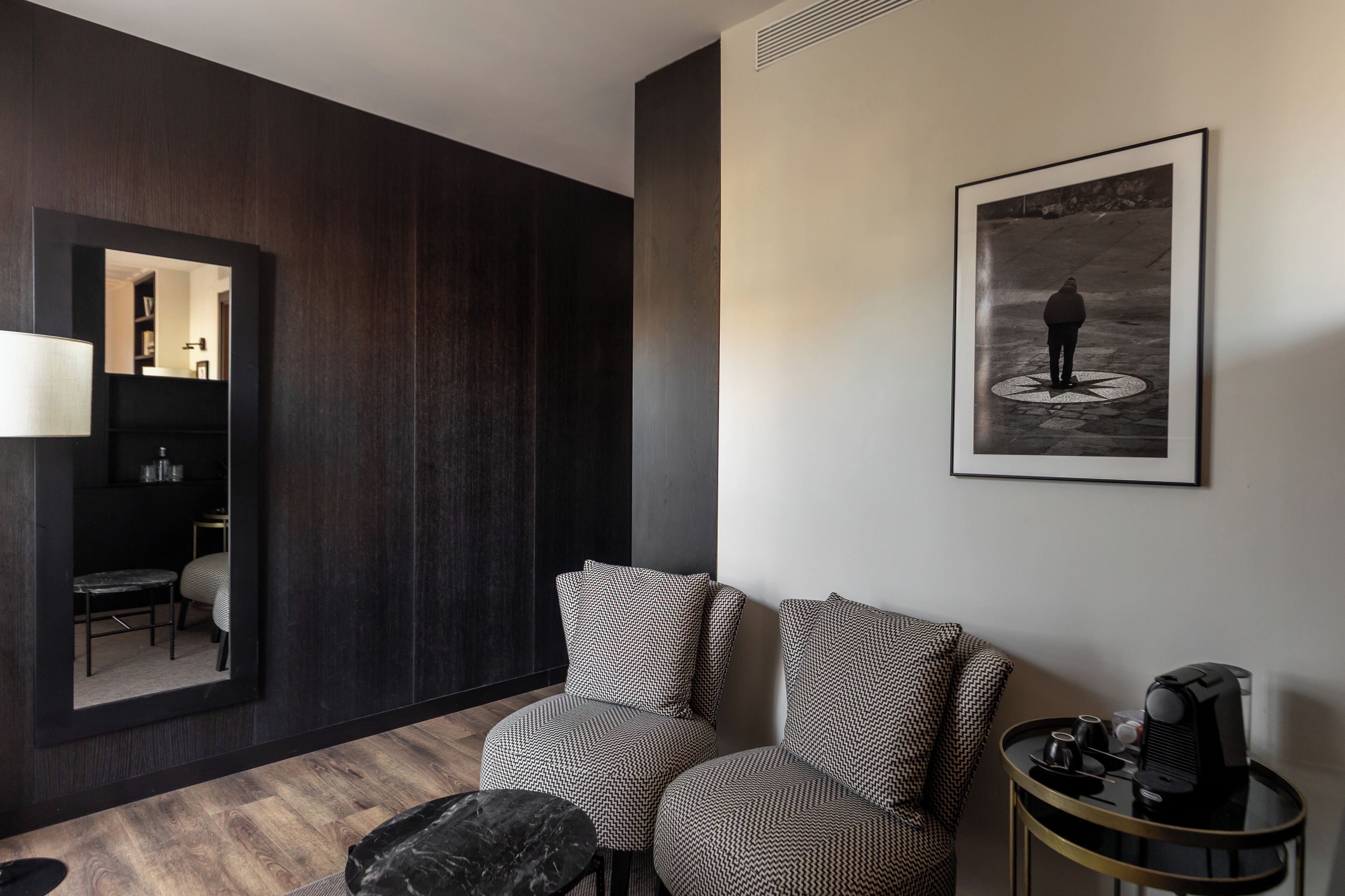 A modern room with dark wood paneling, a large mirror, two patterned armchairs, a small round table, and a framed black and white photograph on the wall.