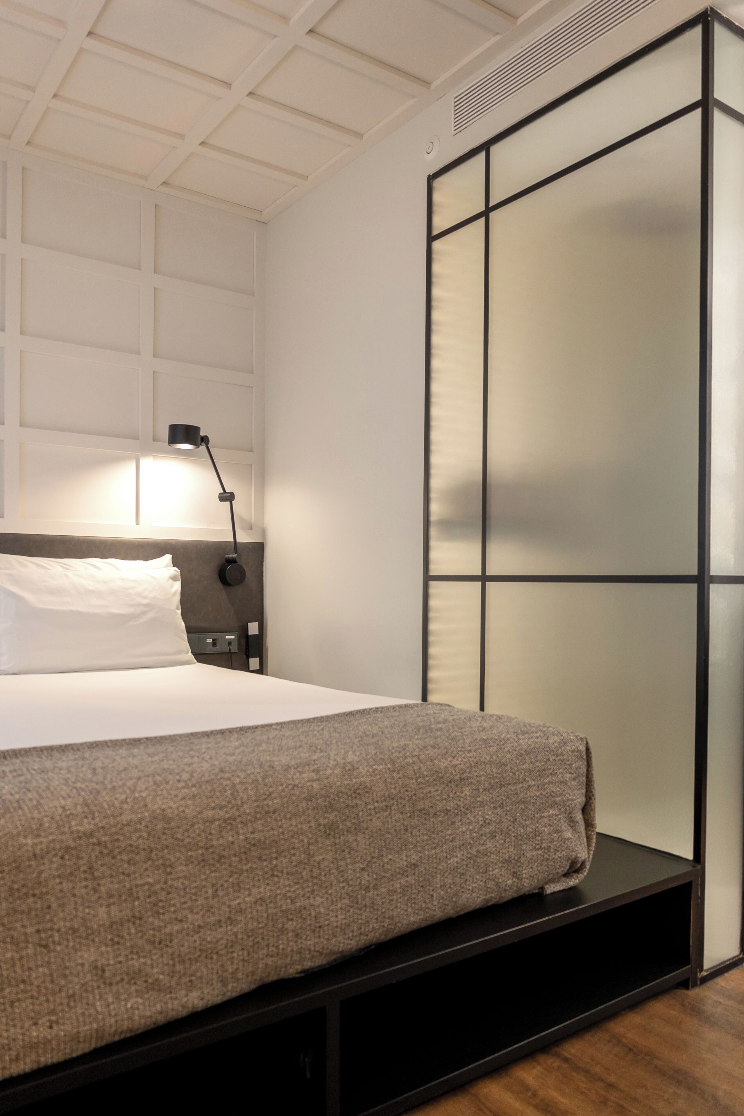 Una cama bien hecha con una almohada blanca y una manta gris, una lámpara de noche y una mesita de noche negra.
